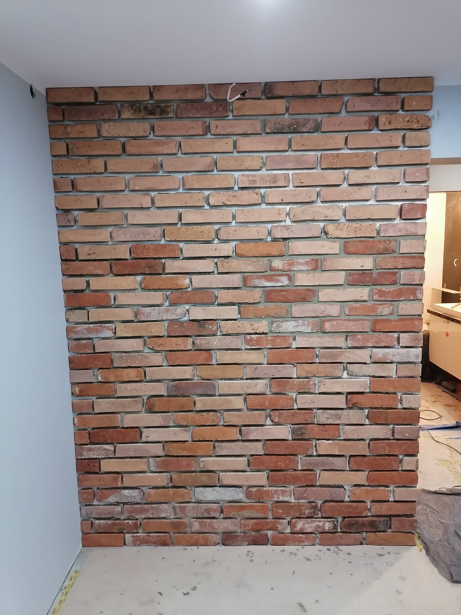 Ściana wykończona czerwoną cegłą klinkierową wewnątrz pomieszczenia, widoczne fragmenty instalacji elektrycznej i niedokończone wykończenie podłogi.