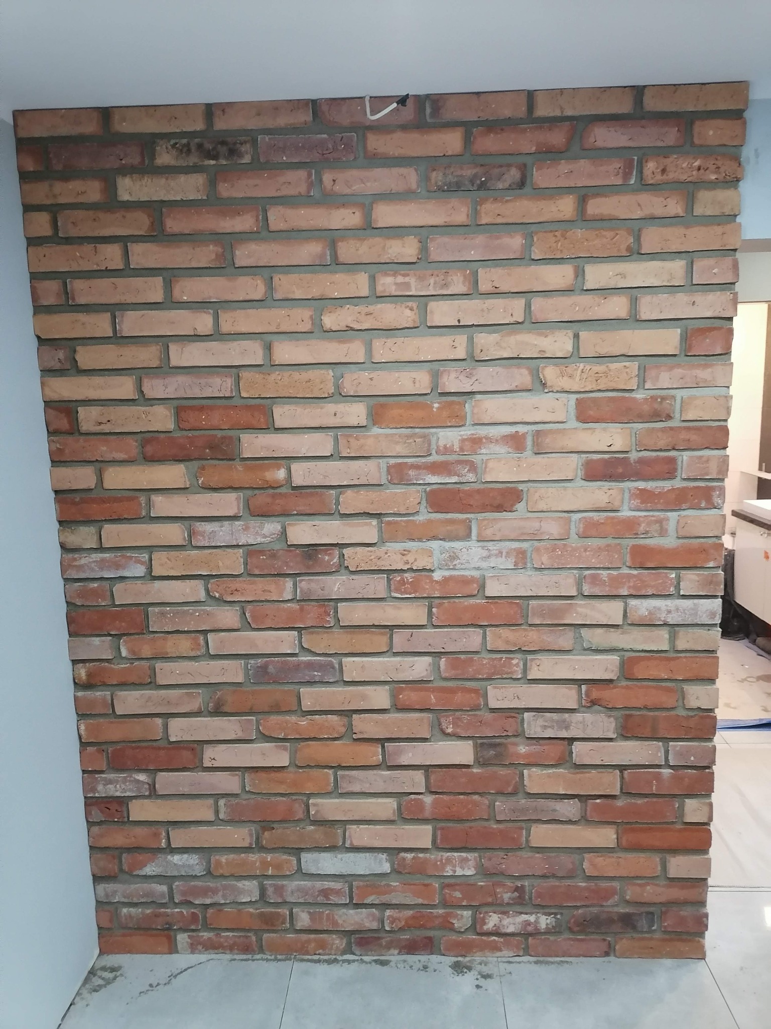Ściana wykończona cegłą klinkierową w odcieniach czerwieni i brązu, z szarą fugą, widoczna w trakcie prac wykończeniowych, z niedoskonałościami na podłodze i ścianach.