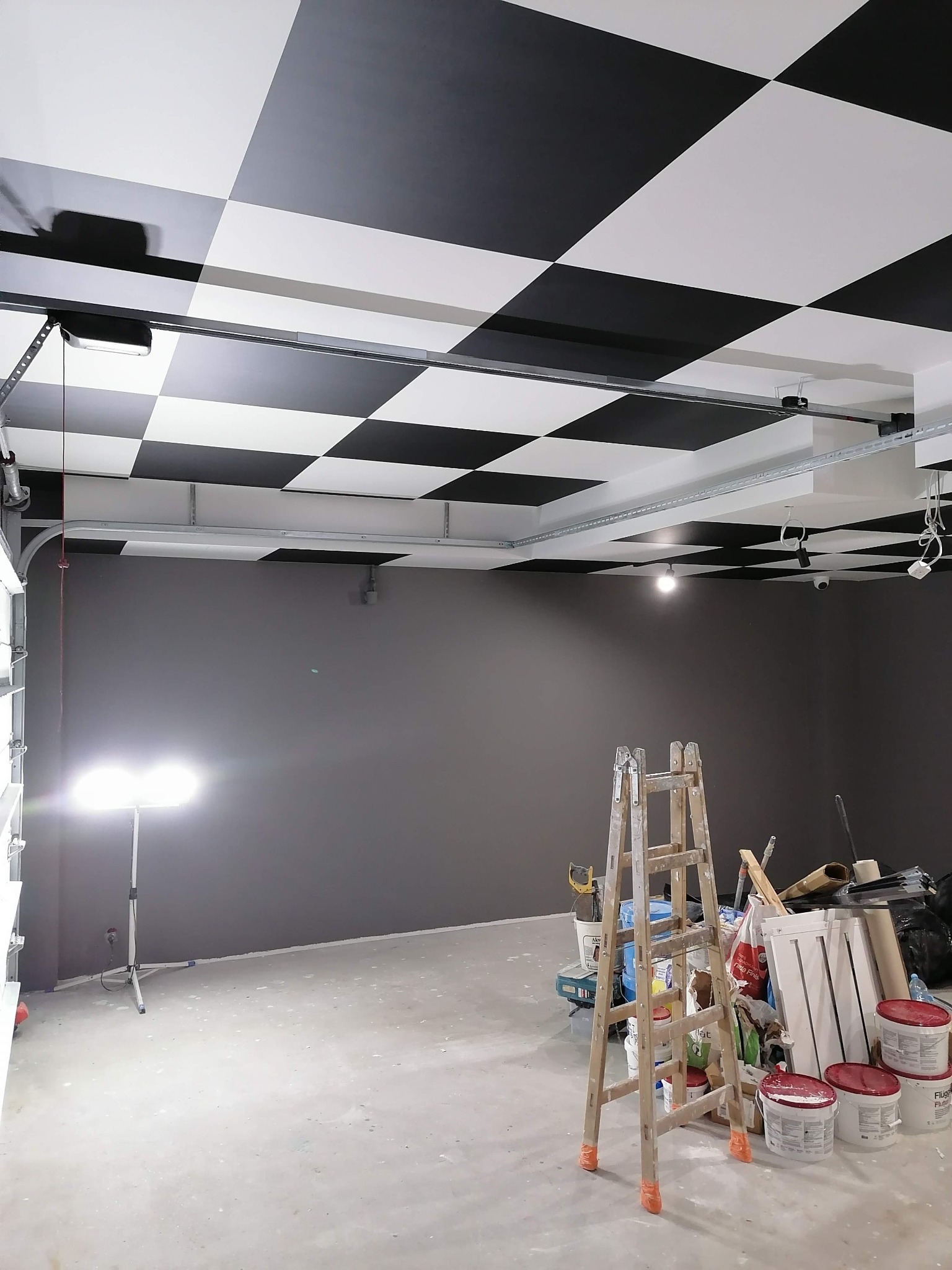 Wnętrze w trakcie prac wykończeniowych: ściana pomalowana na szaro, sufit w czarno-białą szachownicę, drabina i narzędzia malarskie na podłodze, lampa halogenowa oświetla przestrzeń.