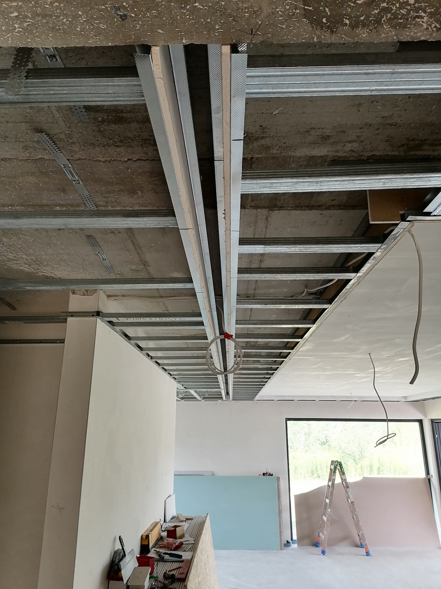 Widok sufitu w trakcie montażu konstrukcji metalowej pod płyty gipsowo-kartonowe, widoczne profile, zawiesia i częściowo zamontowana płyta, w tle ściany i okno z widokiem na zieleń.