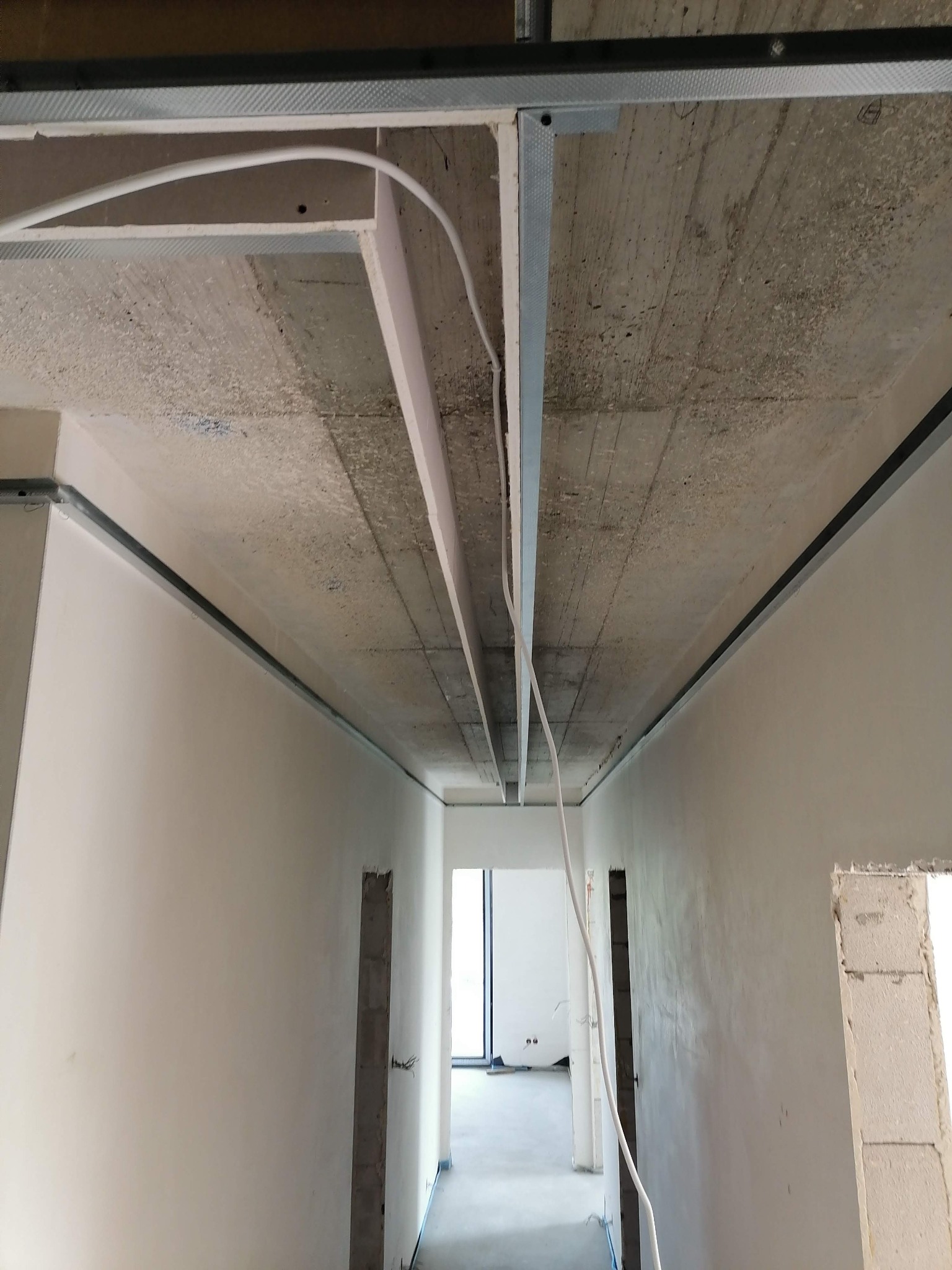 Montaż stelaża sufitu podwieszanego w korytarzu, widoczne profile metalowe, przewody elektryczne i surowy betonowy strop.