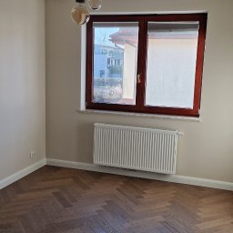 Uni-Mar Marcin Grzelak - Pusty pokój po remoncie z parkietem w jodełkę, świeżo pomalowanymi ścianami w odcieniach beżu i białymi listwami przypodłogowymi, widok na okno z roletą.