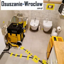 Łazienka w trakcie prac, z widocznym sprzętem do osuszania, w tym żółty osuszacz Trotec Cube+ i instalacja z przewodów zabezpieczonych żółtą taśmą, ściany z częściowo usuniętymi płytkami.