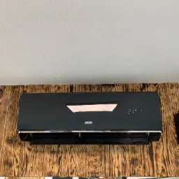 Czarny klimatyzator marki AUX zamontowany na ścianie wyłożonej deskami z wyraźnym rysunkiem drewna, obok widoczny głośnik marki Sven.