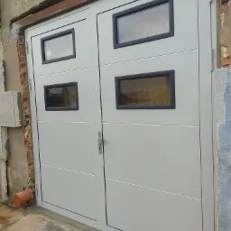 Nowa, dwuskrzydłowa brama garażowa w kolorze szarym z czterema prostokątnymi okienkami z czarną ramą, zamontowana w surowym otworze budynku z widocznymi fragmentami cegieł i betonu.