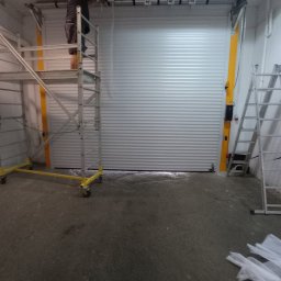 KRAPTECH - Rolety Okna Moskitiery - Montaż białej bramy garażowej rolowanej; pracownik na rusztowaniu reguluje mechanizm, drabina oparta o ścianę, widoczne elementy montażowe i folie ochronne na podłodze.