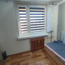 Wnętrze pokoju z oknem wyposażonym w rolety dzień-noc, obok okna firanka, pod oknem grzejnik, w rogu pokoju widoczny fragment łóżka.