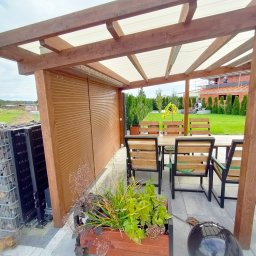 KRAPTECH - Rolety Okna Moskitiery - Zadaszona drewniana altana z brązowymi listwami, chroniąca przestrzeń do siedzenia z drewnianym stołem i krzesłami, obok widoczna konstrukcja z kamieniami w metalowej siatce.