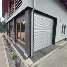 KRAPTECH - Rolety Okna Moskitiery - Zamknięta, srebrna brama segmentowa garażowa z poziomymi przetłoczeniami, zamontowana w szarej elewacji budynku z oknami o drewnianych ramach, widok z zewnątrz.