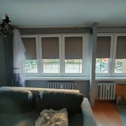 KRAPTECH - Rolety Okna Moskitiery - Wnętrze pokoju z trzema oknami z szarymi roletami zwiniętymi do połowy, widoczne grzejniki pod oknami i fragment sofy z przodu.