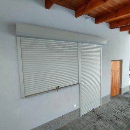 KRAPTECH - Rolety Okna Moskitiery - Szara roleta zewnętrzna częściowo opuszczona na oknie, obok drewniane drzwi wejściowe, widoczne elementy drewnianej konstrukcji dachu i fragment ośnieżonego ogrodu w tle.