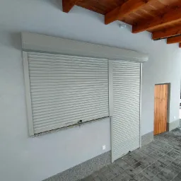 Szara roleta zewnętrzna częściowo opuszczona na oknie, obok drewniane drzwi wejściowe, widoczne elementy drewnianej konstrukcji dachu i fragment ośnieżonego ogrodu w tle.