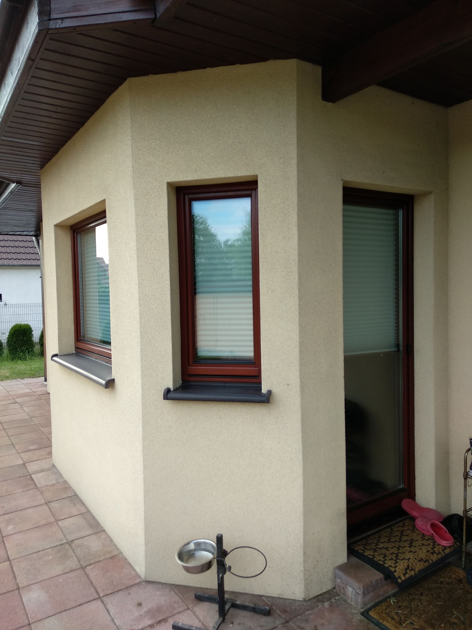 Narożnik domu z dwoma oknami drewnianymi z żaluzjami plisowanymi i parapetem aluminiowym, widoczna miska dla psa na stojaku, brązowa stolarka okienna.