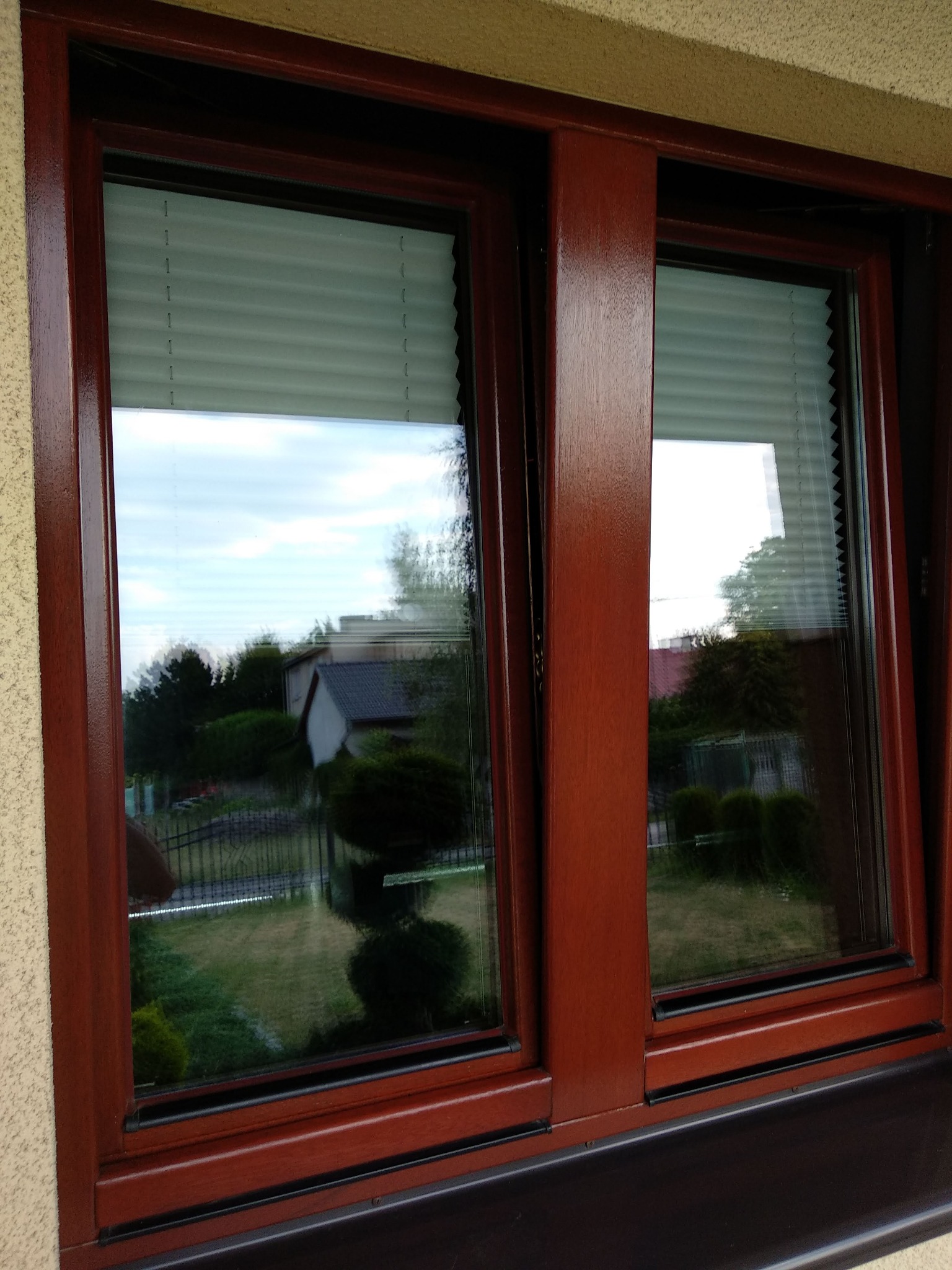 Dwu-skrzydłowe okno z brązową ramą, widok na ogród przysłonięty plisowaną żaluzją, ciemny parapet zewnętrzny.