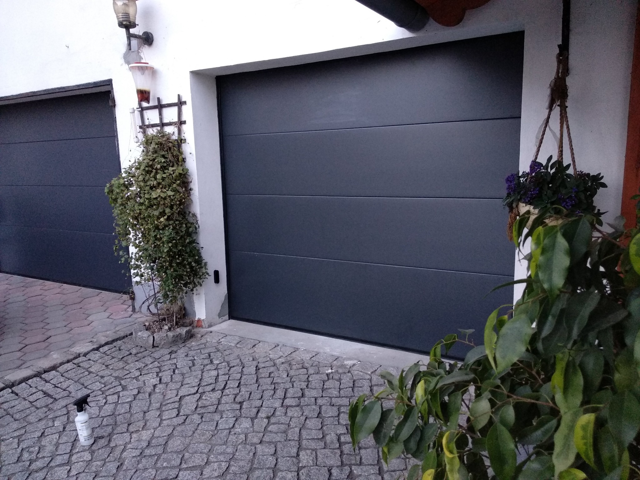 Nowoczesna, grafitowa brama garażowa segmentowa z poziomymi przetłoczeniami, widoczna z zewnątrz na tle kostki brukowej, z fragmentem drugiej bramy po lewej stronie i wiszącą doniczką z kwiatami po...