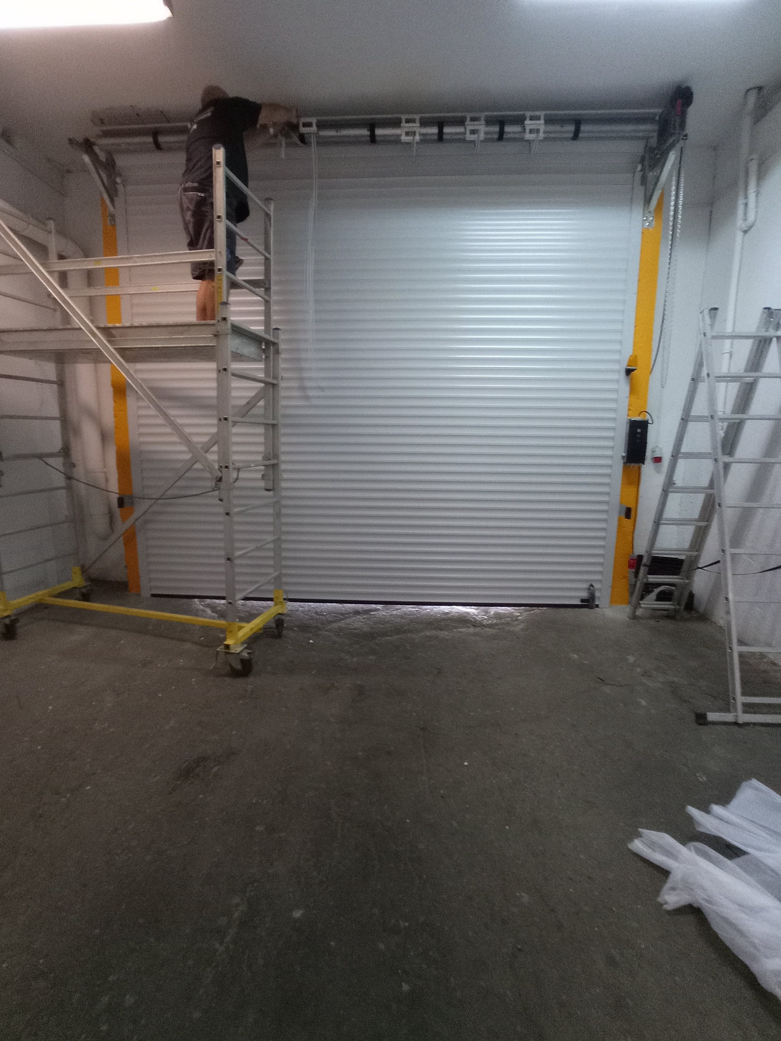 Montaż białej bramy garażowej rolowanej; pracownik na rusztowaniu reguluje mechanizm, drabina oparta o ścianę, widoczne elementy montażowe i folie ochronne na podłodze.