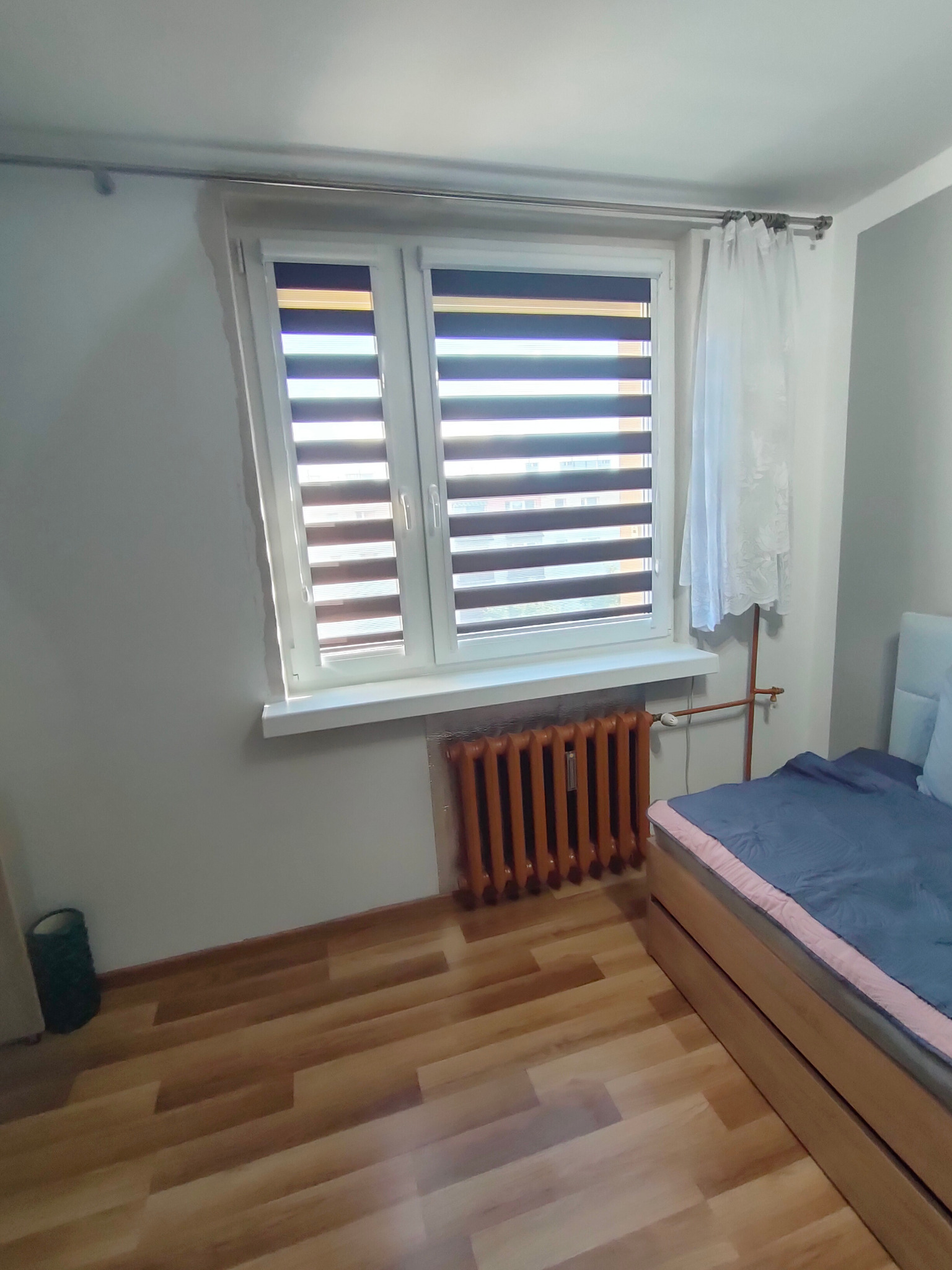 Wnętrze pokoju z oknem wyposażonym w rolety dzień-noc, obok okna firanka, pod oknem grzejnik, w rogu pokoju widoczny fragment łóżka.