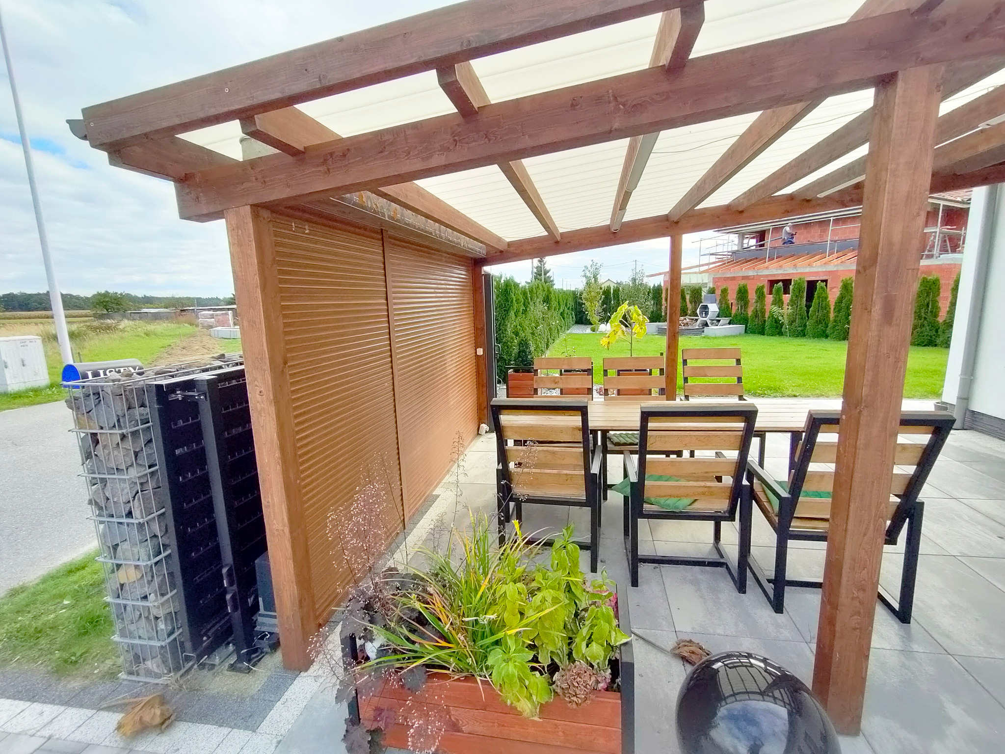 Zadaszona drewniana altana z brązowymi listwami, chroniąca przestrzeń do siedzenia z drewnianym stołem i krzesłami, obok widoczna konstrukcja z kamieniami w metalowej siatce.