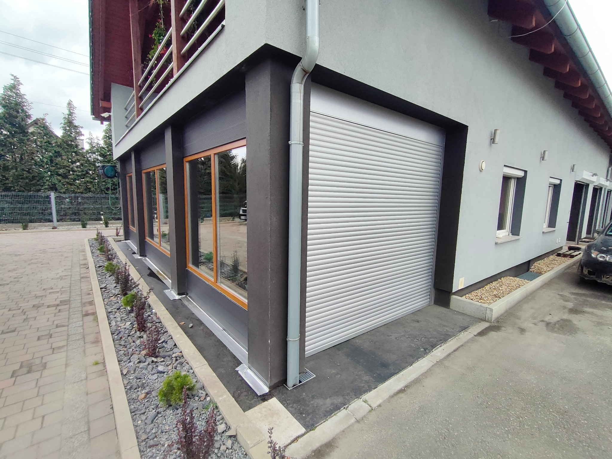 Zamknięta, srebrna brama segmentowa garażowa z poziomymi przetłoczeniami, zamontowana w szarej elewacji budynku z oknami o drewnianych ramach, widok z zewnątrz.