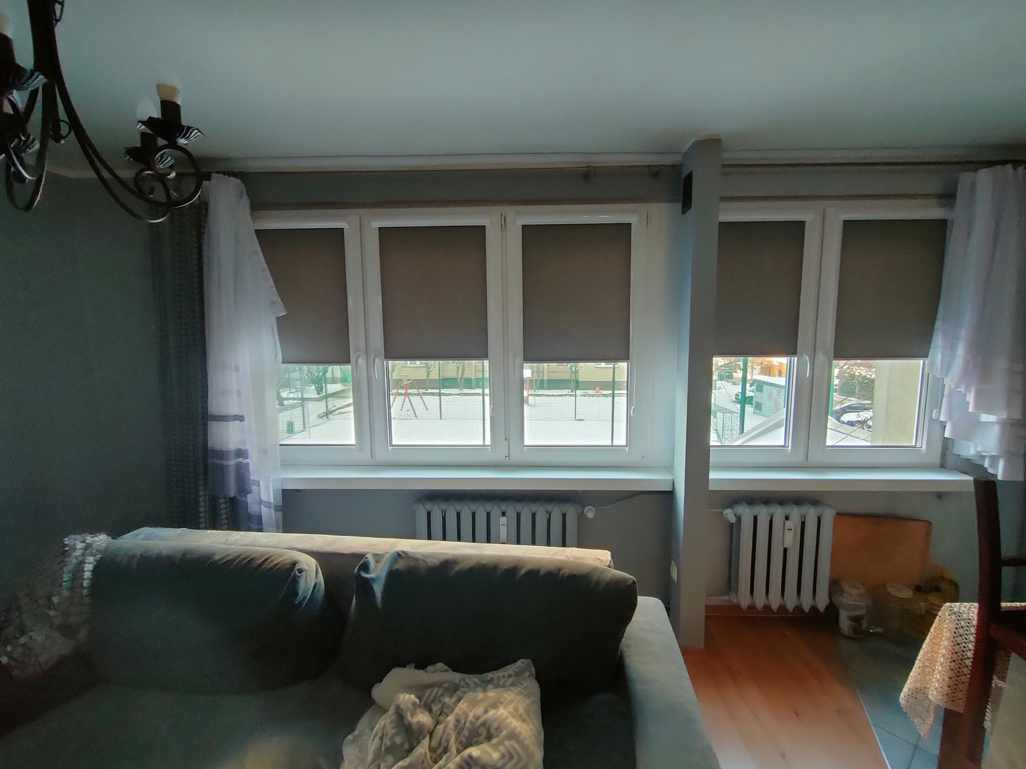 Wnętrze pokoju z trzema oknami z szarymi roletami zwiniętymi do połowy, widoczne grzejniki pod oknami i fragment sofy z przodu.