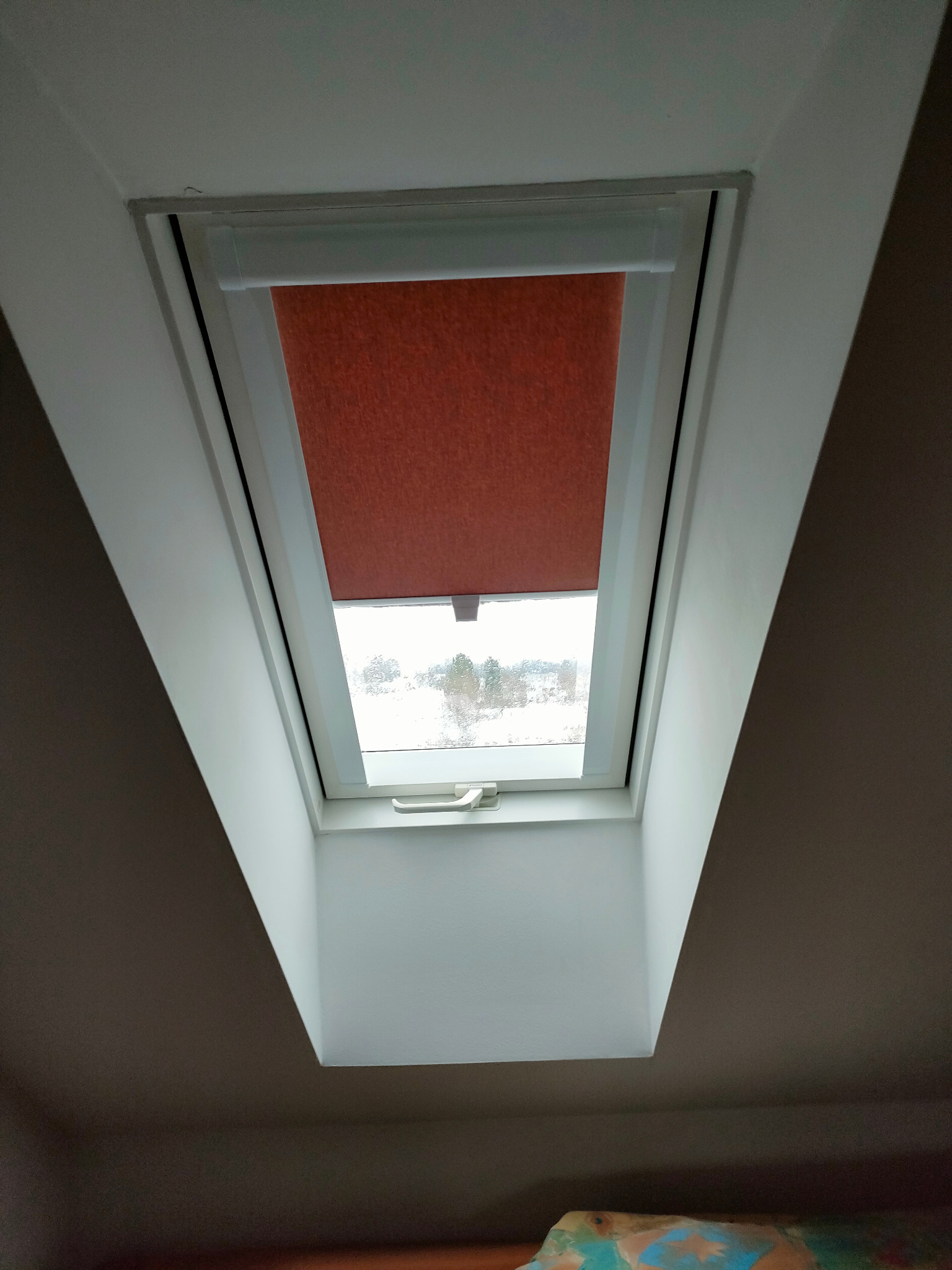 Okno dachowe z częściowo zasłoniętą, pomarańczową roletą, widok na zimowy krajobraz za oknem.