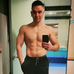 Mężczyzna z widoczną muskulaturą, robiący selfie w szatni, w tle umywalka i lustro.