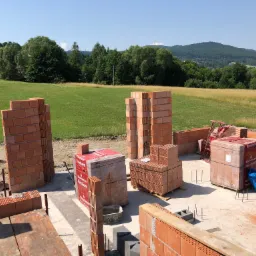 Budowa domu z czerwonej cegły Porotherm na tle zielonego pola i zalesionych wzgórz w słoneczny dzień.