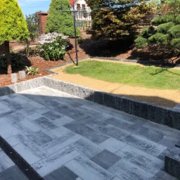 Szare kostki brukowe ułożone na posesji, z odpływem liniowym i obrzeżami, otoczone zielenią i korą.