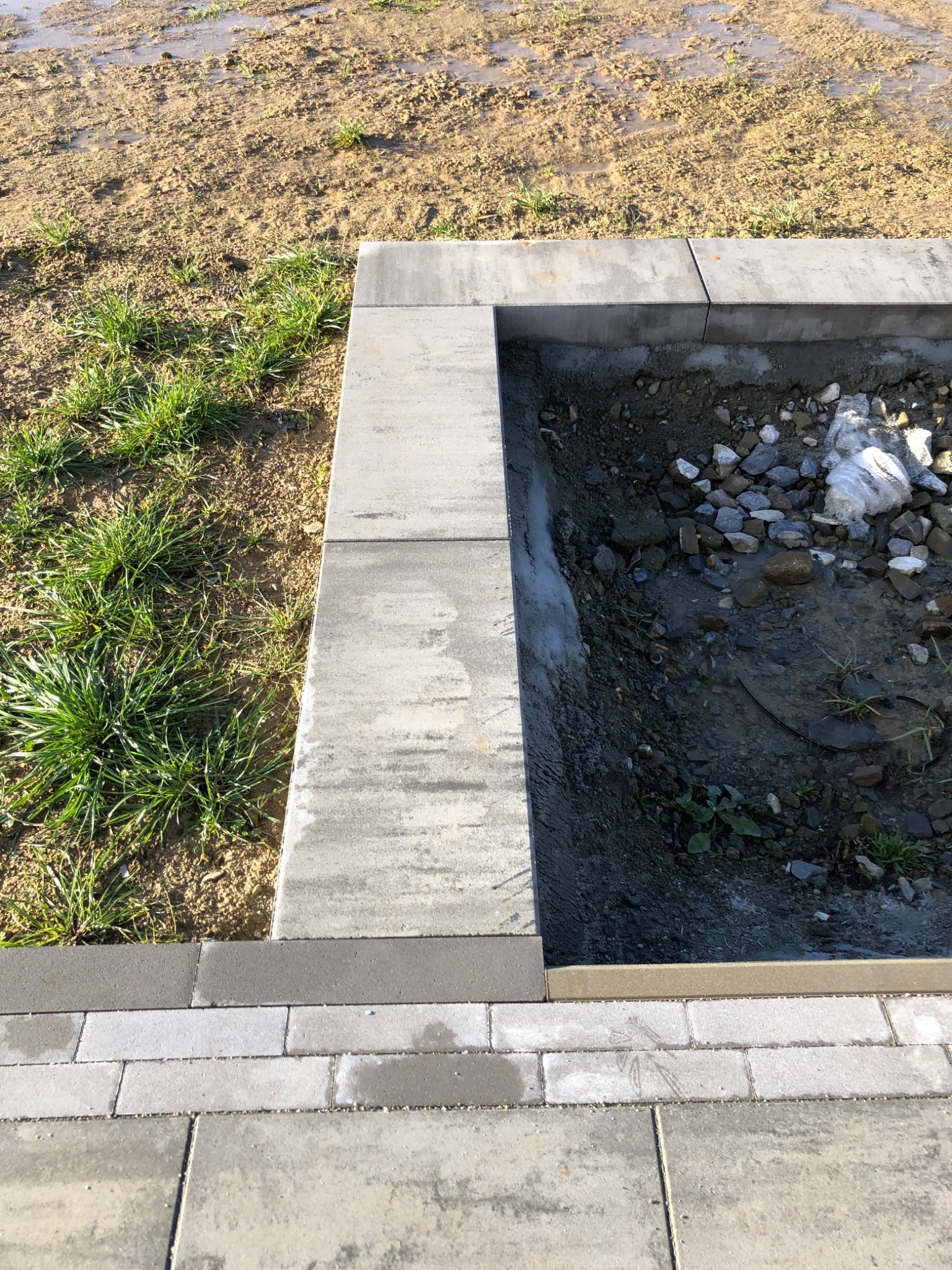 Przygotowanie podłoża pod obrzeże z kostki brukowej: widoczne ułożone obrzeże, wykop wypełniony kamieniami i gruzem, oraz mokra ziemia w tle.