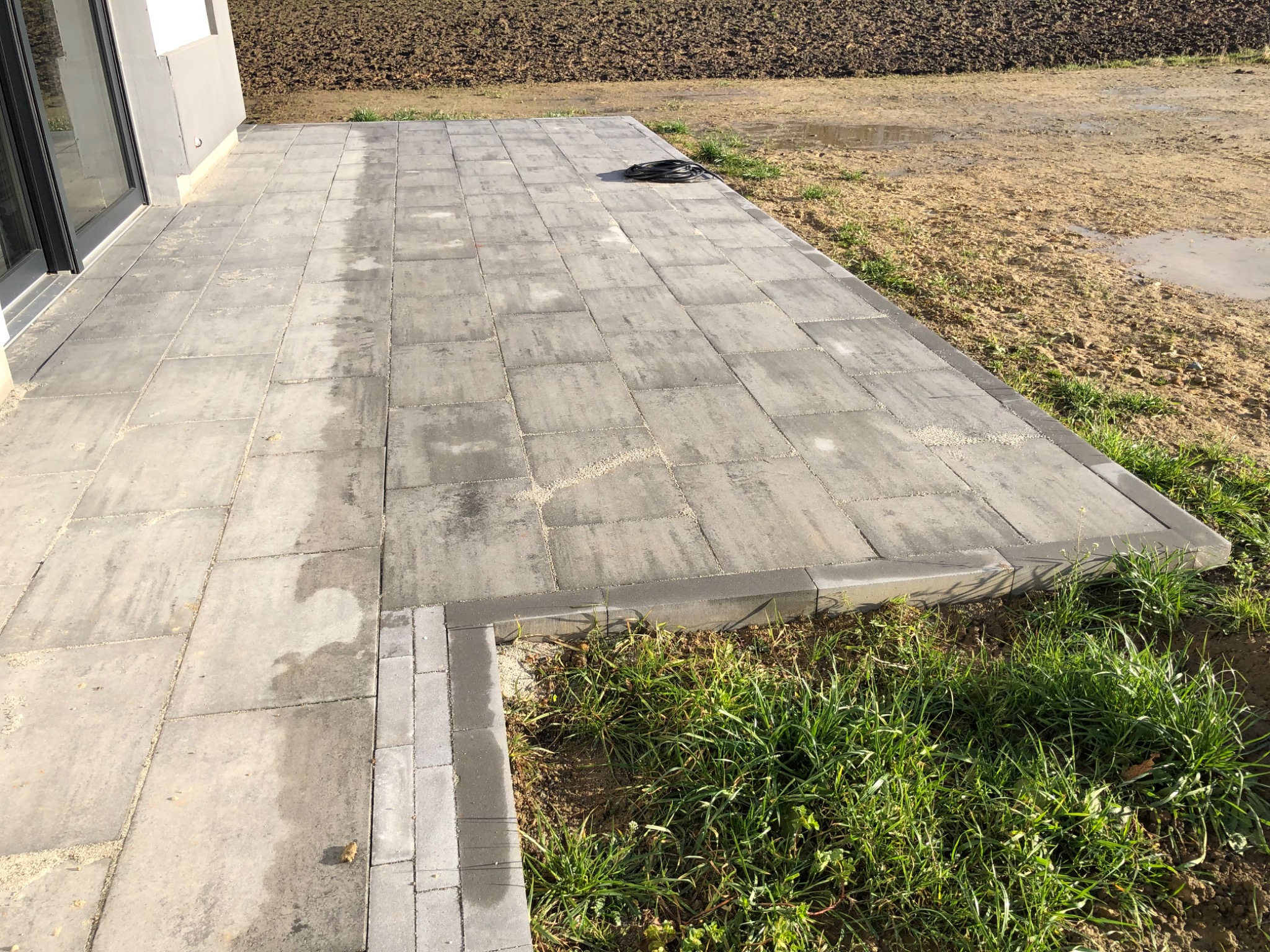 Nowoczesny taras z szarej kostki brukowej, obrzeża z drobniejszej kostki, widoczne ślady wilgoci i kabel na powierzchni, tło z ziemią i młodą trawą.