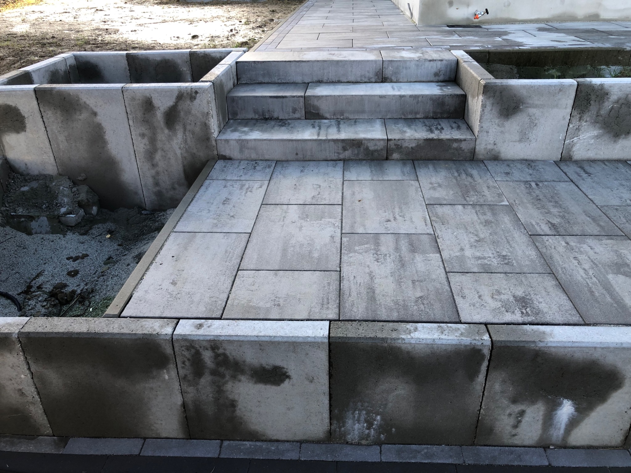 Wykończona ścieżka z szarych płyt betonowych prowadząca do schodów i podwyższonych rabat z betonowych bloczków, widoczne ślady wilgoci na powierzchni bloczków.