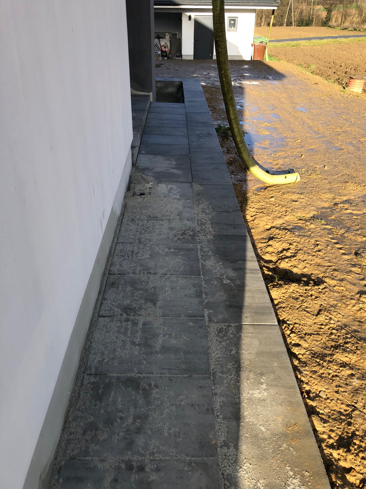 Świeżo ułożona, szara kostka brukowa przy białej ścianie budynku, z widoczną rurą odprowadzającą wodę na błotniste podłoże.