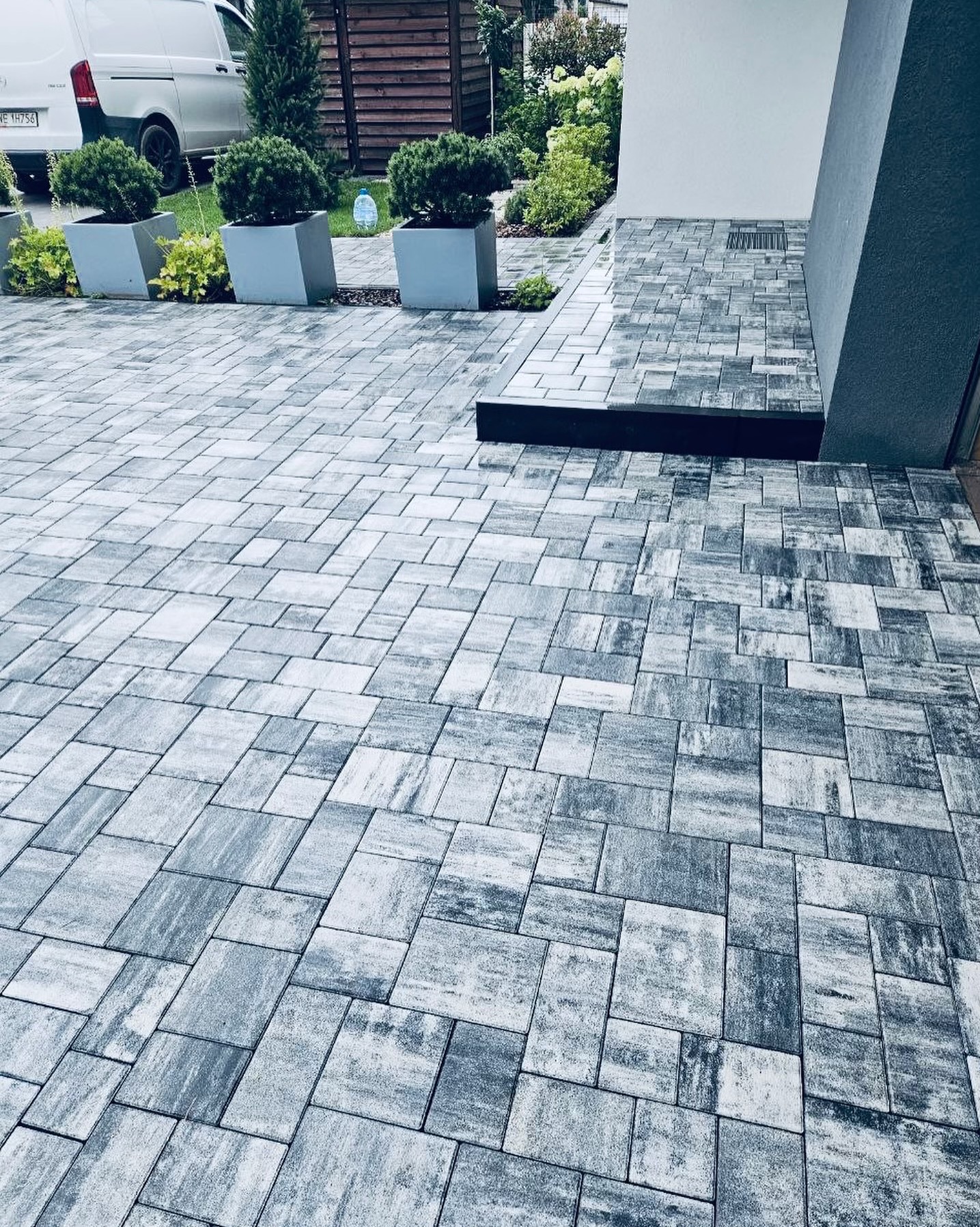 Ułożona szara kostka brukowa o zróżnicowanym wzorze, z donicami z roślinami i fragmentem elewacji budynku w tle.