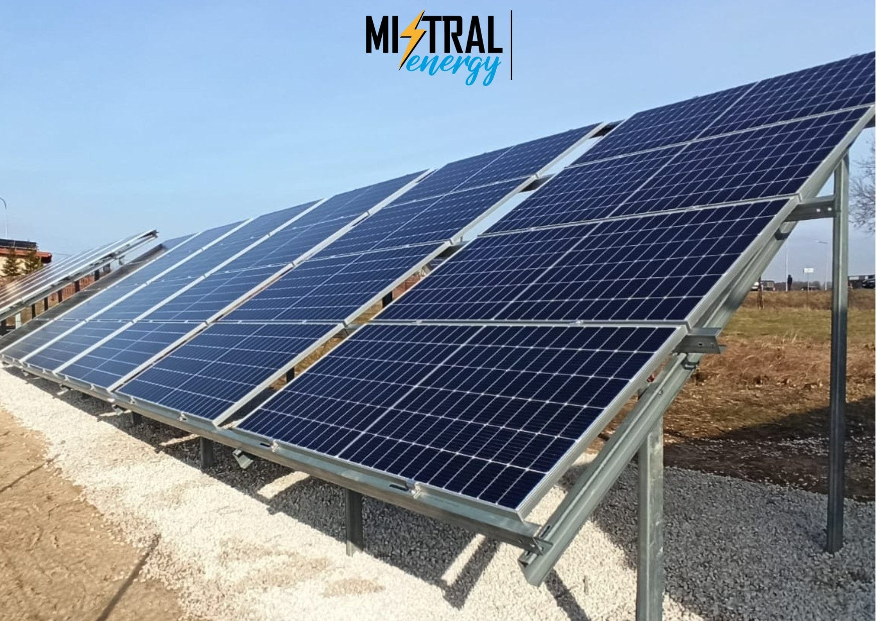 Rząd paneli słonecznych zamontowanych na metalowej konstrukcji, widoczna marka Mistral Energy w górnym rogu, tło to błękitne niebo i odległy krajobraz.
