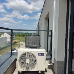 Zewnętrzna jednostka klimatyzacji marki AUX zamontowana na balkonie w nowoczesnym budynku mieszkalnym, widok na okolicę z błękitnym niebem i chmurami.