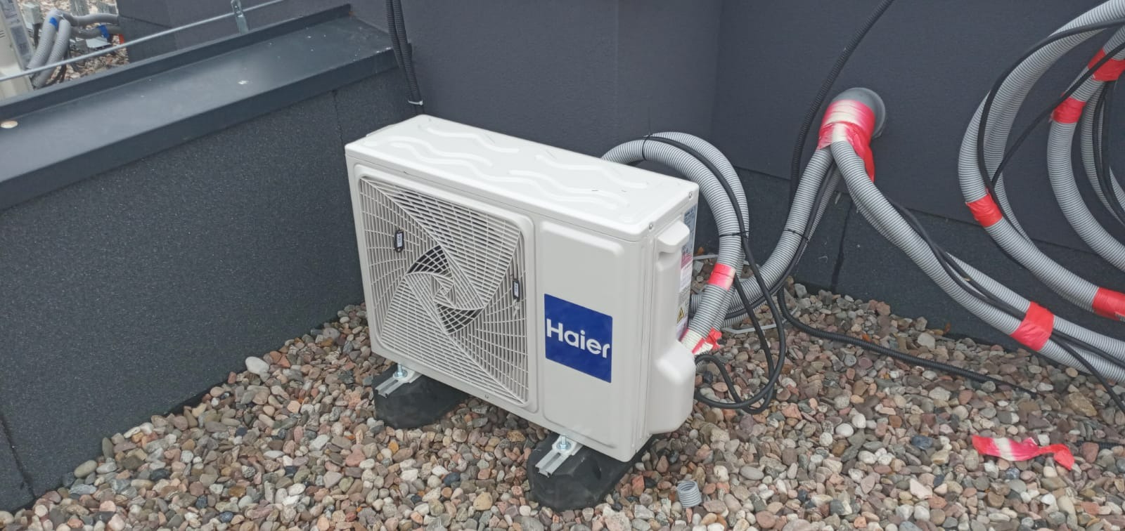 Zewnętrzna jednostka klimatyzacji Haier zainstalowana na dachu pokrytym żwirem, widoczne przewody w peszlach z czerwoną taśmą izolacyjną.