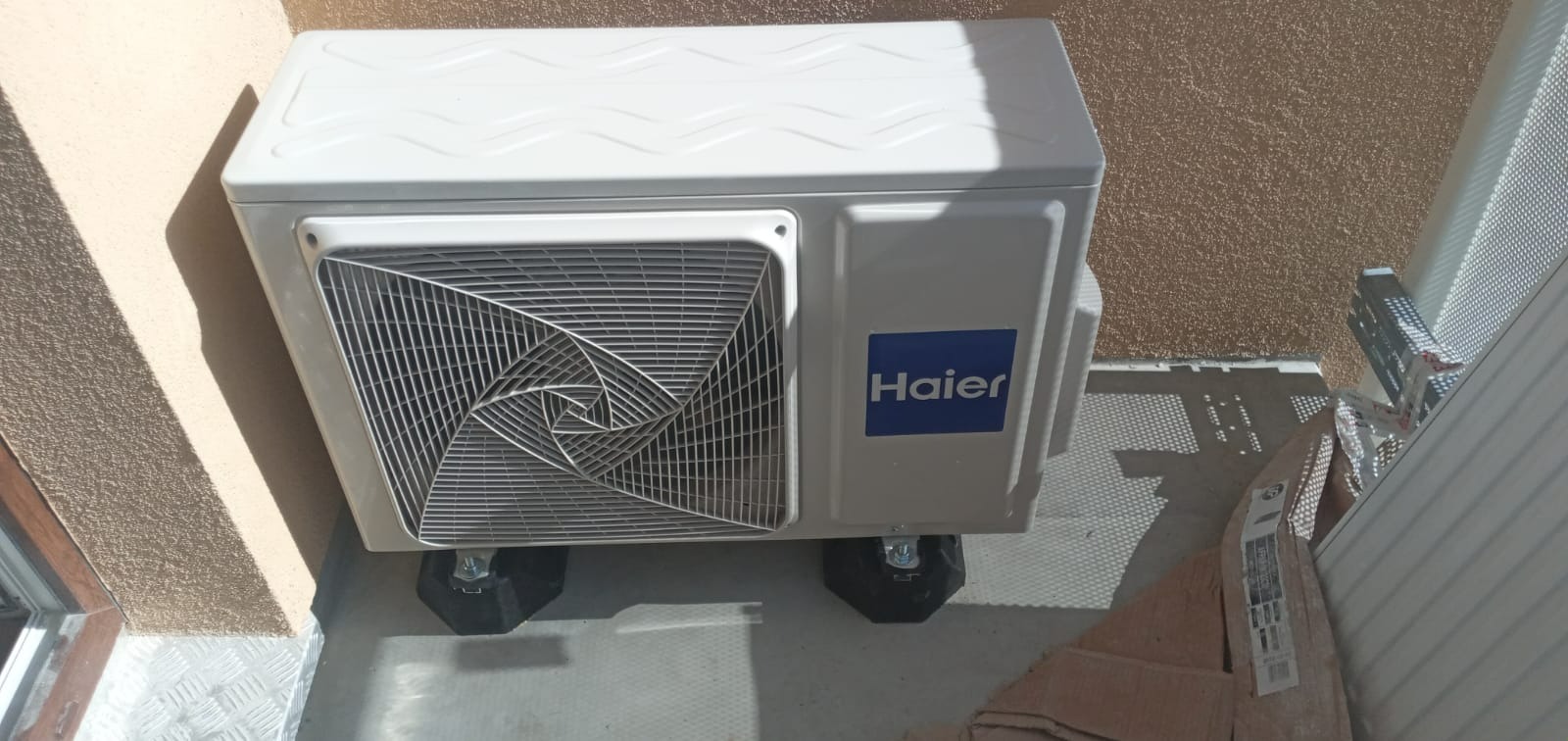 Zewnętrzna jednostka klimatyzacji Haier zamontowana na czarnych podkładkach antywibracyjnych na balkonie, widoczna kratka wentylacyjna i logo producenta.