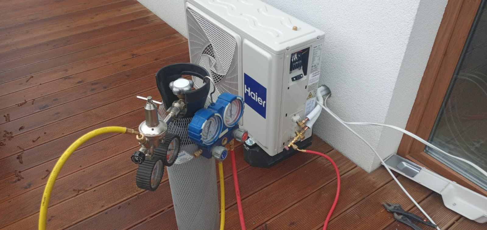 Zewnętrzna jednostka klimatyzacji Haier zamontowana na elewacji domu, podłączona do butli z manometrami na drewnianym tarasie podczas serwisu.