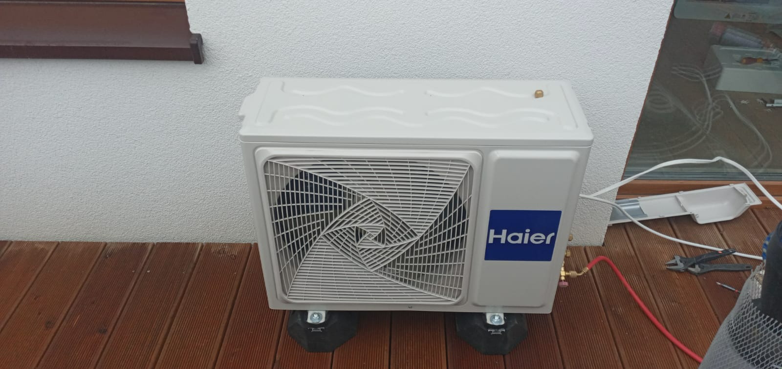 Biała jednostka zewnętrzna klimatyzacji Haier umieszczona na czarnych podkładkach antywibracyjnych na drewnianym tarasie, widoczne narzędzia i przewody instalacyjne.