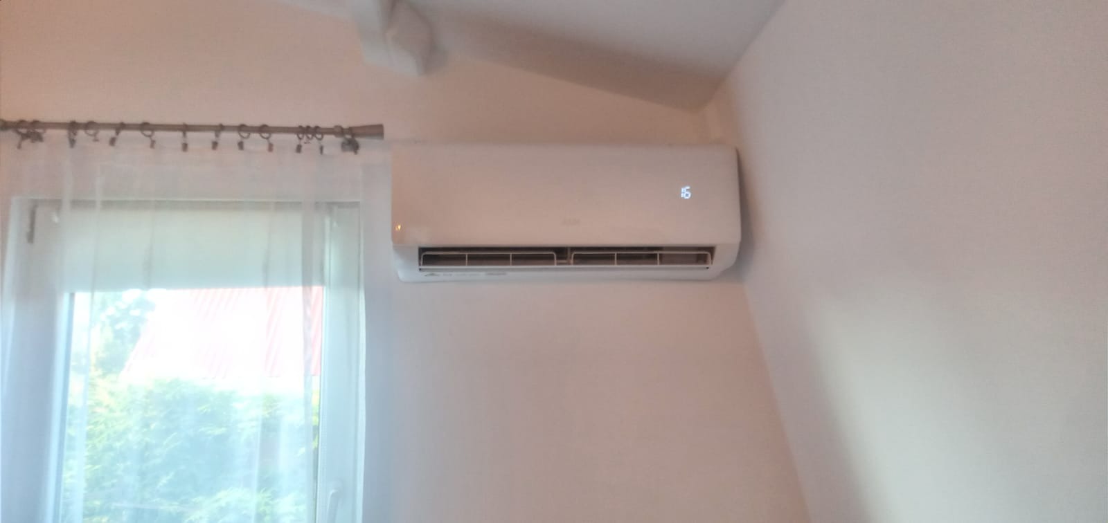 Biały klimatyzator ścienny w pokoju z oknem zasłoniętym przezroczystą firanką, ustawiony na temperaturę 16 stopni Celsjusza.