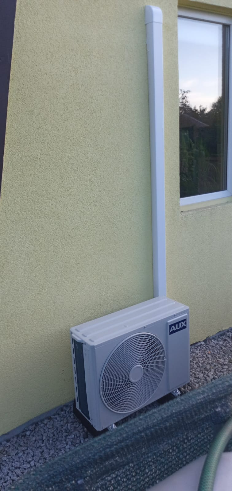 Zewnętrzna jednostka klimatyzacji AUX zamontowana przy żółtej, teksturowanej ścianie domu, z widoczną białą rurą odprowadzającą skropliny i oknem w tle.