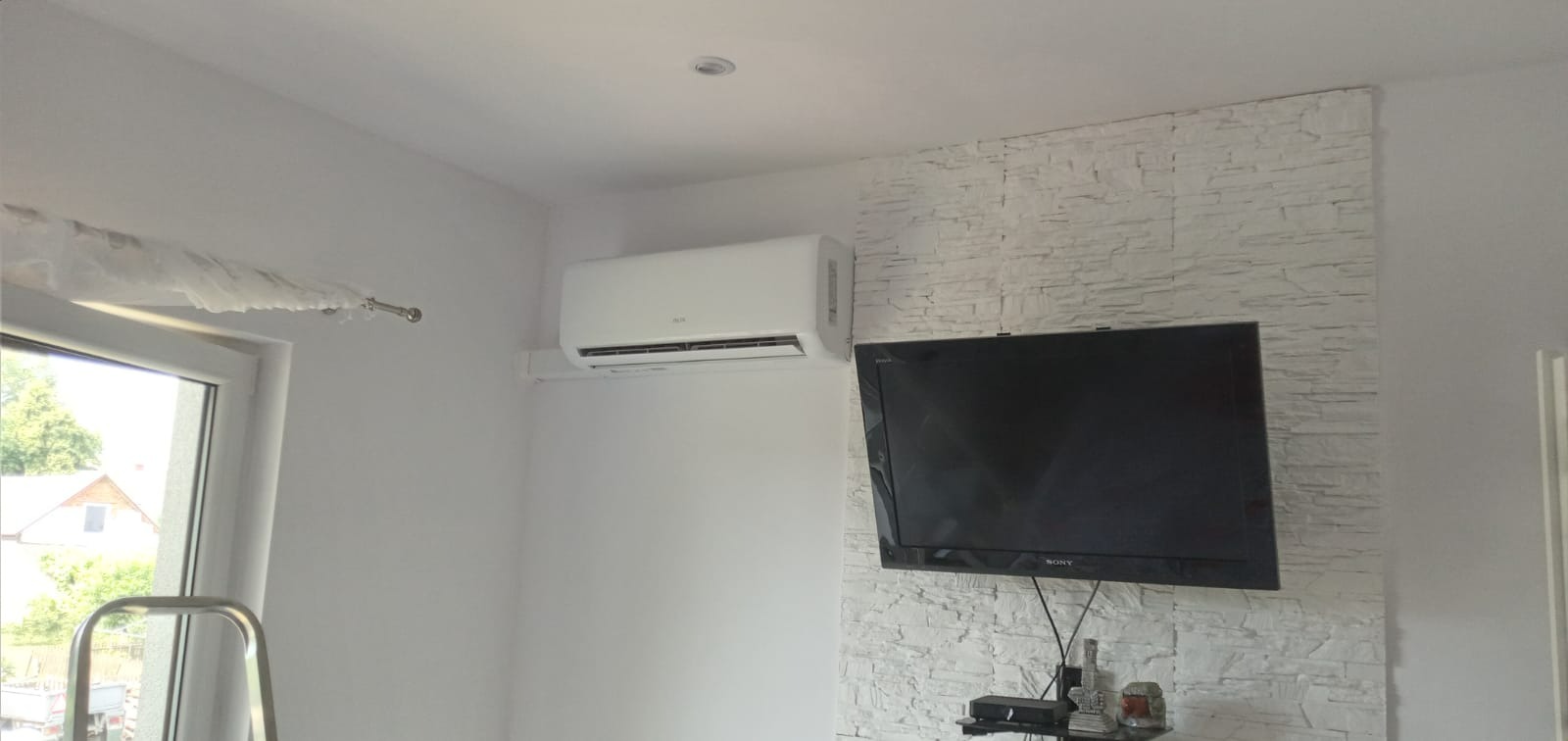 Biały klimatyzator ścienny zamontowany w pokoju z telewizorem na dekoracyjnej ścianie z białej cegły.