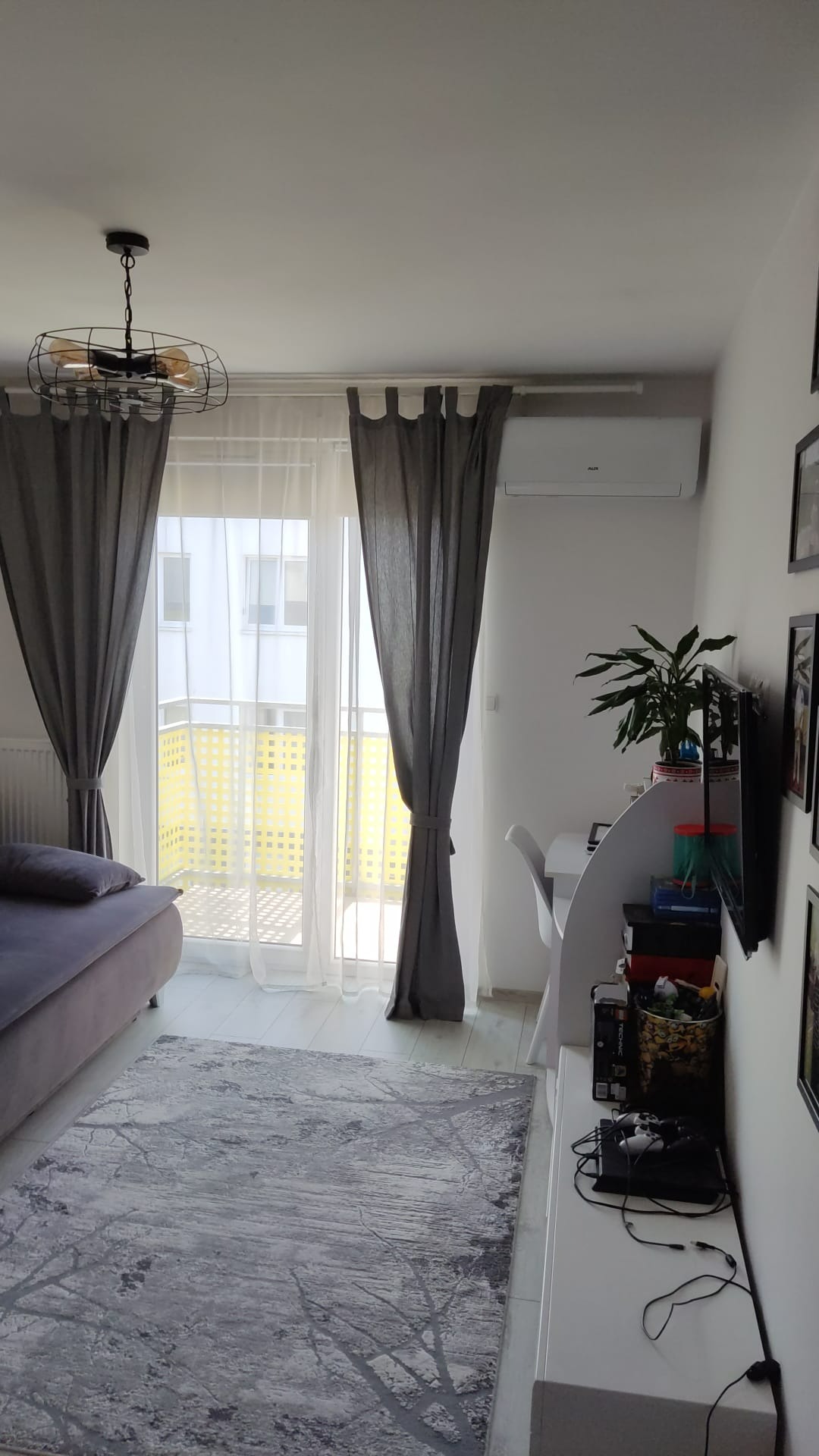 Jasne wnętrze pokoju z zamontowanym klimatyzatorem AUX nad oknem balkonowym z szarymi zasłonami.