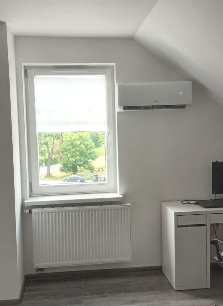 Biały klimatyzator zamontowany na ścianie w pokoju z oknem, widoczny grzejnik pod oknem i biurko z komputerem.