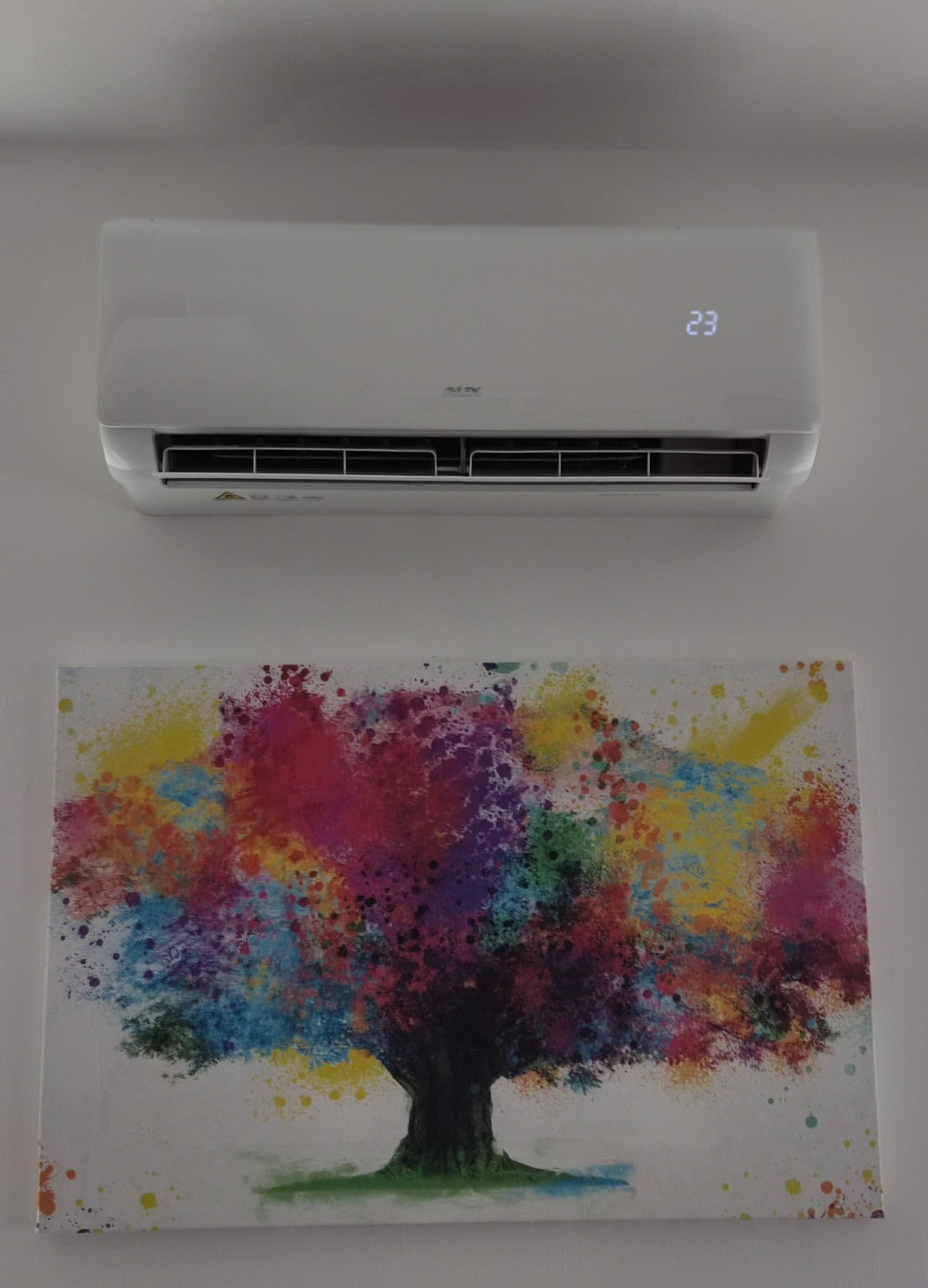 Biały klimatyzator ścienny z wyświetlaną temperaturą 23 stopnie, zawieszony nad kolorowym obrazem drzewa na białej ścianie.