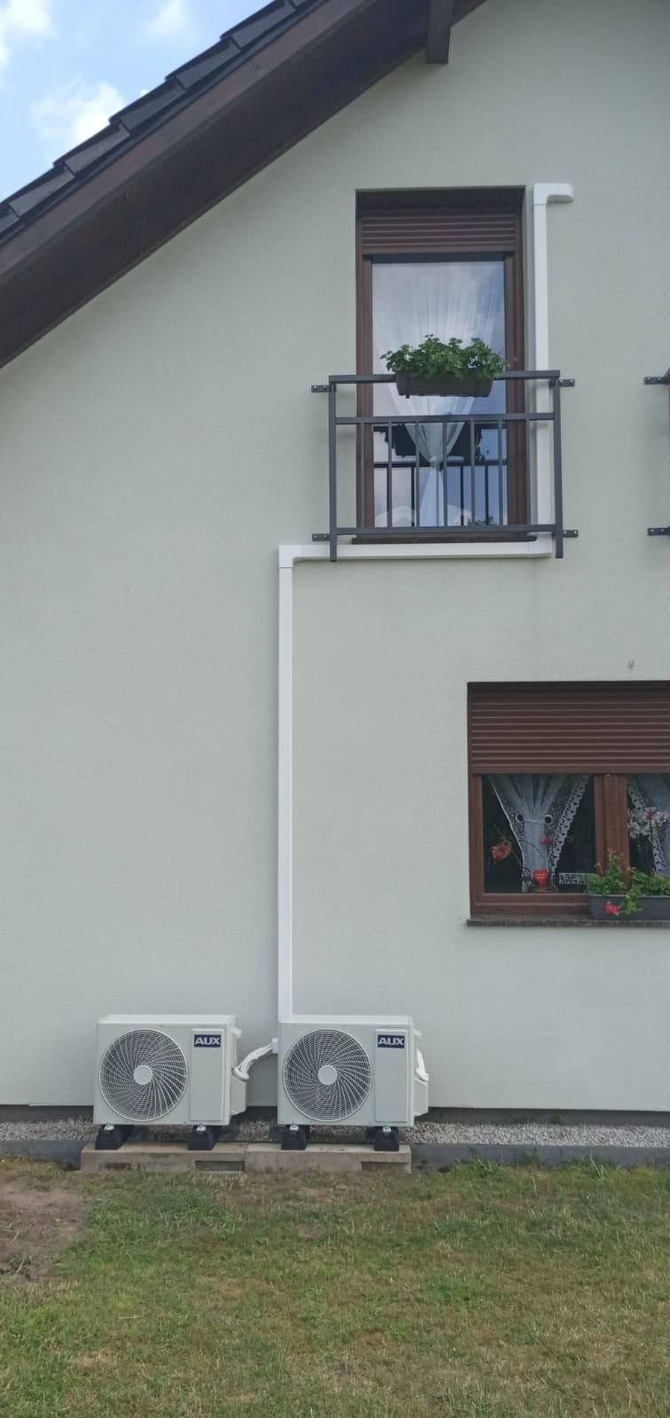 Dwa zewnętrzne agregaty klimatyzacji marki AUX zamontowane na betonowych podstawach przy ścianie domu jednorodzinnego z oknami z brązowymi roletami i balkonem z kwiatami.