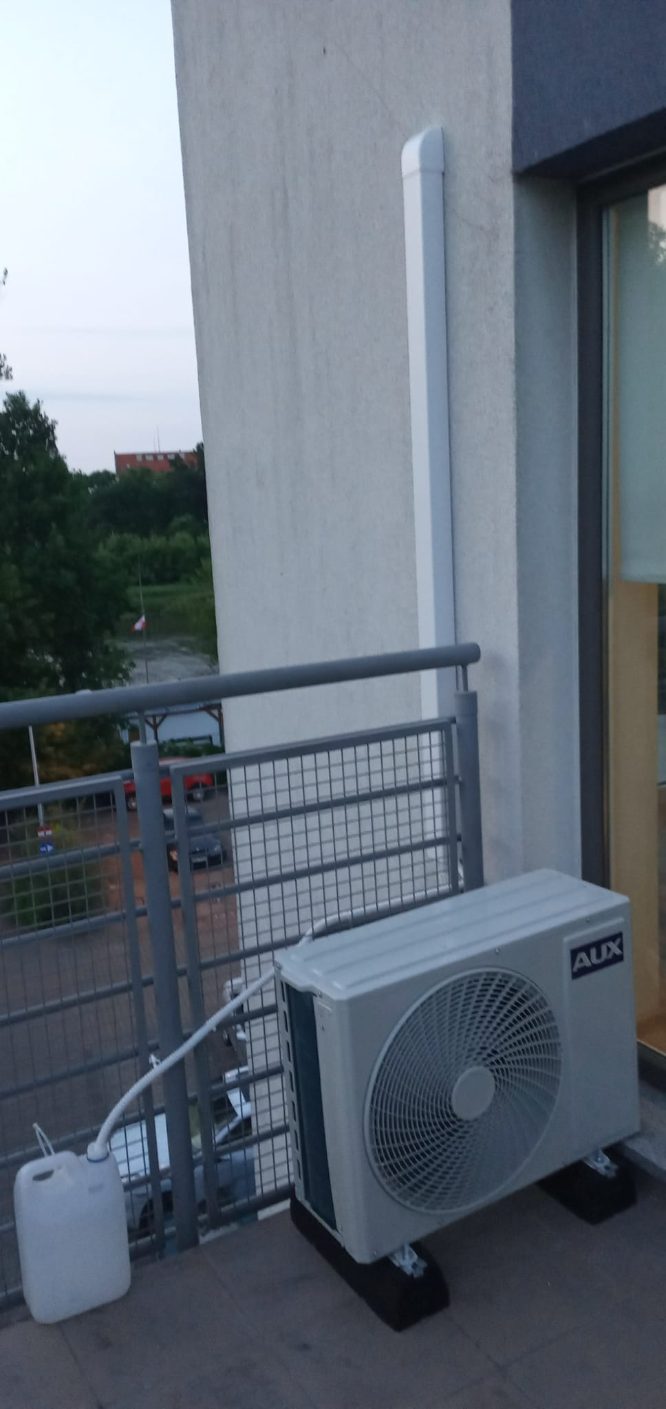 Jednostka zewnętrzna klimatyzatora AUX na balkonie z podłączonym pojemnikiem na skropliny, widoczny odpływ.