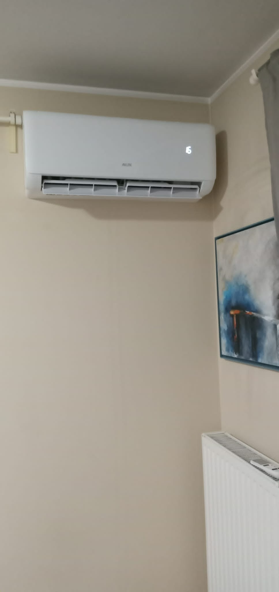Biała jednostka klimatyzacyjna marki AUX zamontowana na beżowej ścianie w pomieszczeniu, obok obrazu w niebieskiej ramie i białego grzejnika.