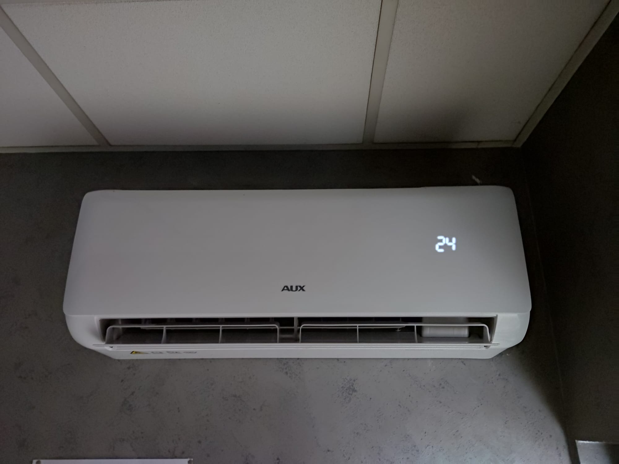 Zamontowany biały klimatyzator AUX z wyświetlaczem temperatury 24 stopnie Celsjusza na szarej ścianie w pomieszczeniu z podwieszanym sufitem.