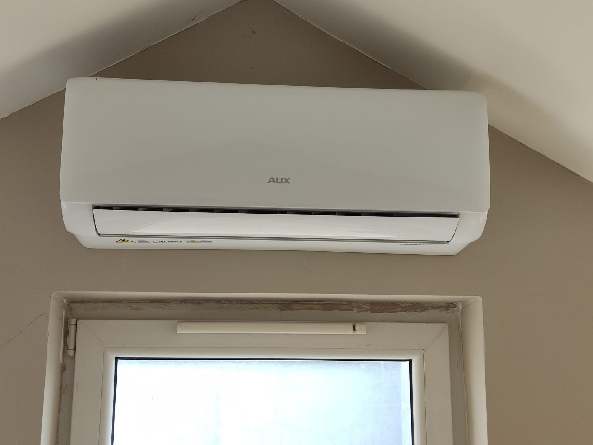 Biała klimatyzacja marki AUX zamontowana nad oknem w domu, widoczne uszczelki okienne wymagające odświeżenia.